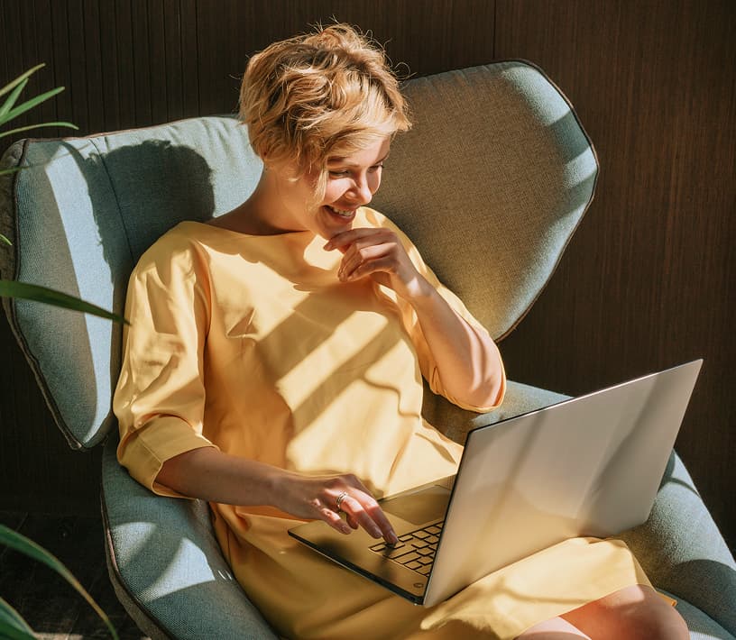 A smiling woman in a yellow dress uses a laptop while sitting in a blue armchair.