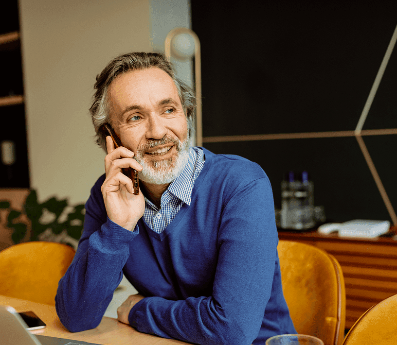 A smiling bearded man in a blue sweater talks on a smartphone.